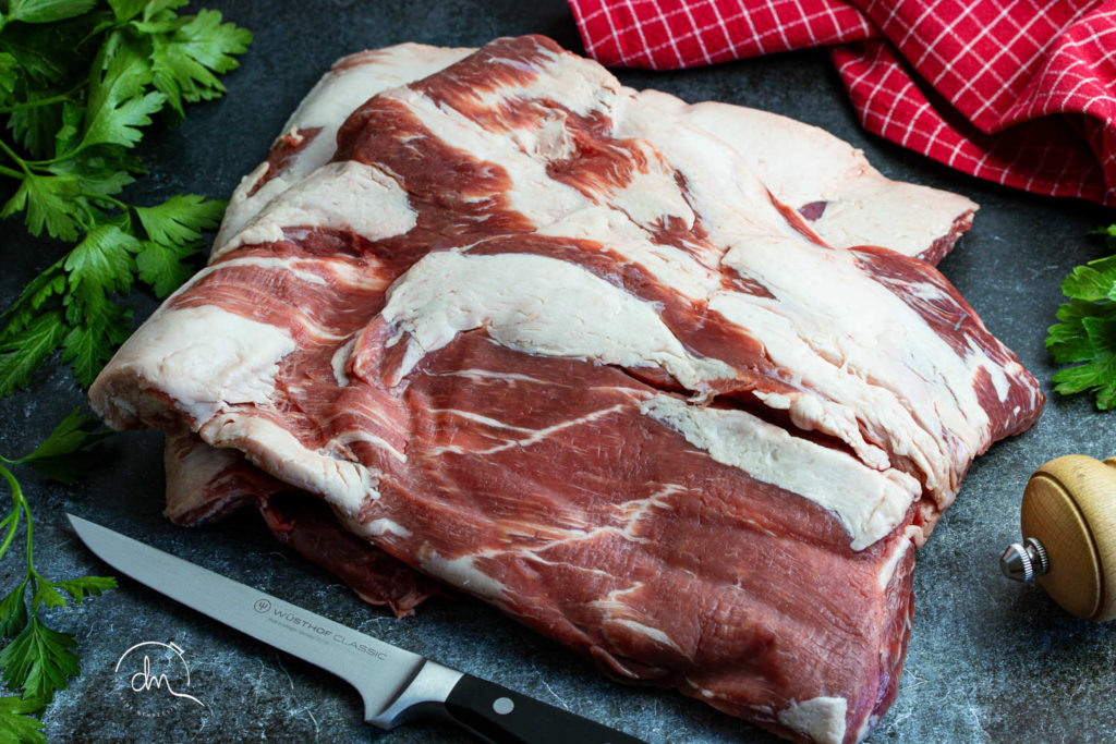 Poitrine 3D de porc noir de Bigorre AOP crue, posée sur une planche avec persil et torchon à carreaux rouges