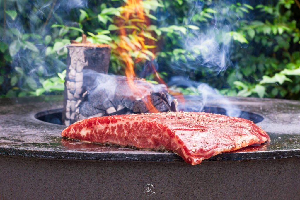 Bavette Wagyu assaisonnée posée sur un brasero plancha en feu avec fumée et verdure en arrière-plan